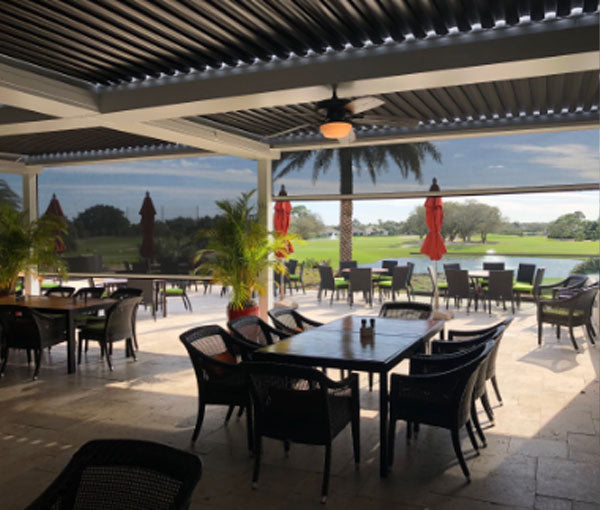 A covered outdoor dining terrace with a louvered roof, set with tables and chairs.