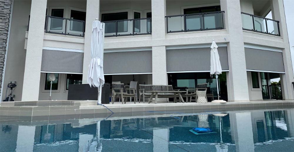 A swimming pool in front of a two-storey home, with motorized screens lowered across the covered patio and the balcony above it.