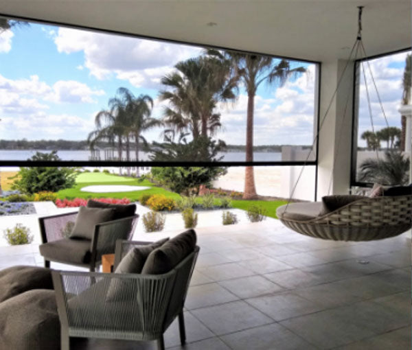 A covered patio with hanging chairs and a motorized screen lowered on the open side, looking out over a lawn and water.