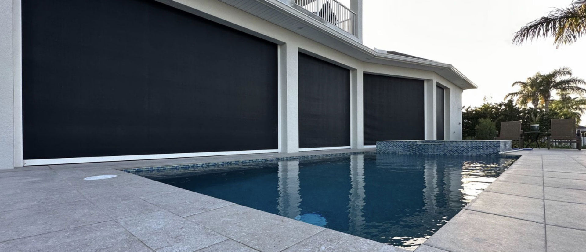 Dark motorized solar screens lowered across a long covered patio beside a swimming pool.